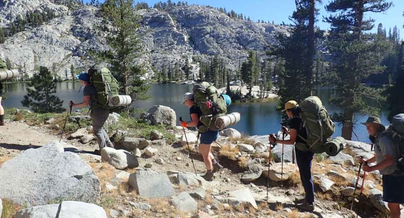 a group of people carrying backpacks hike past a body of water with mountains in the background